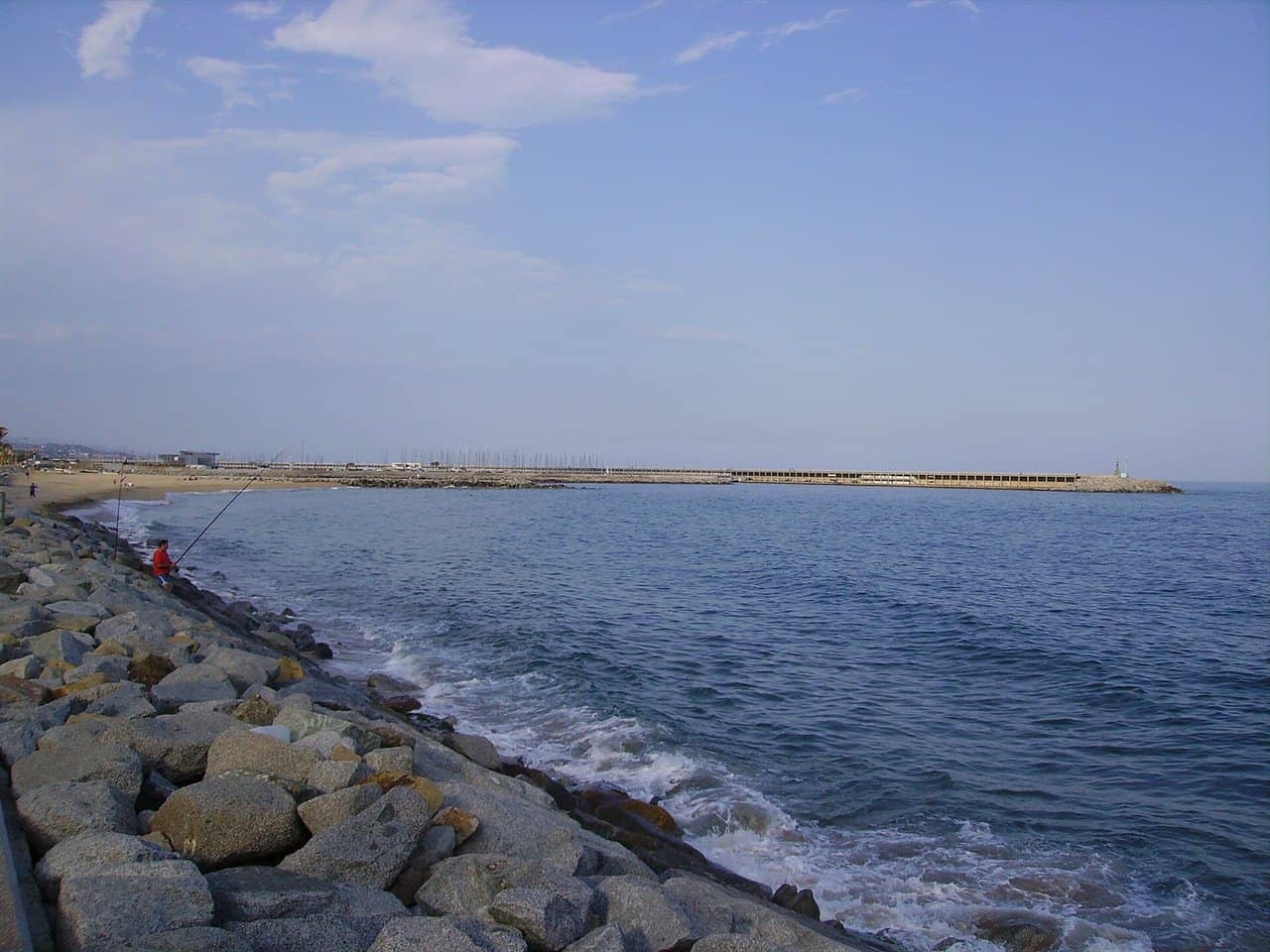 Orilla rocosa con un pescador y, al fondo, el espigón del puerto de Mataró