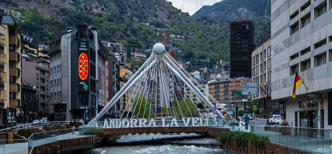 Pont amb el rètol Andorra la Vella sobre el riu al centre de la ciutat, amb les muntanyes al fons