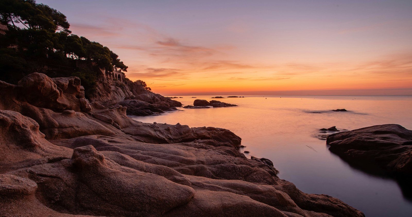 Sunset over the rocky Costa Brava shore: pines on the stones and a calm sea