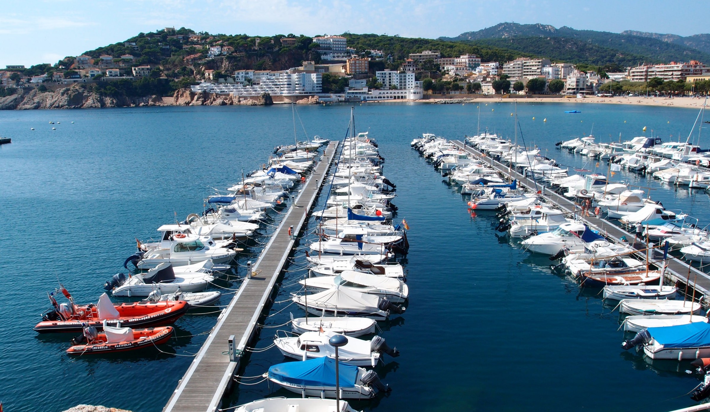 El puerto deportivo de Sant Feliu de Guíxols: las embarcaciones en los pantalanes, la playa y el cabo sobre la bahía